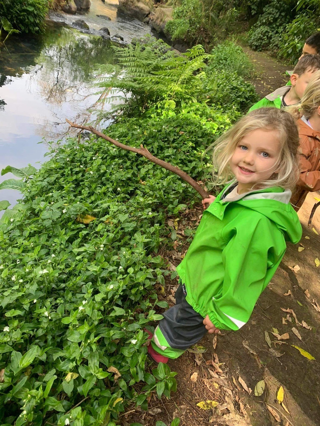 Photo of child in green coat holding a stick near a stream